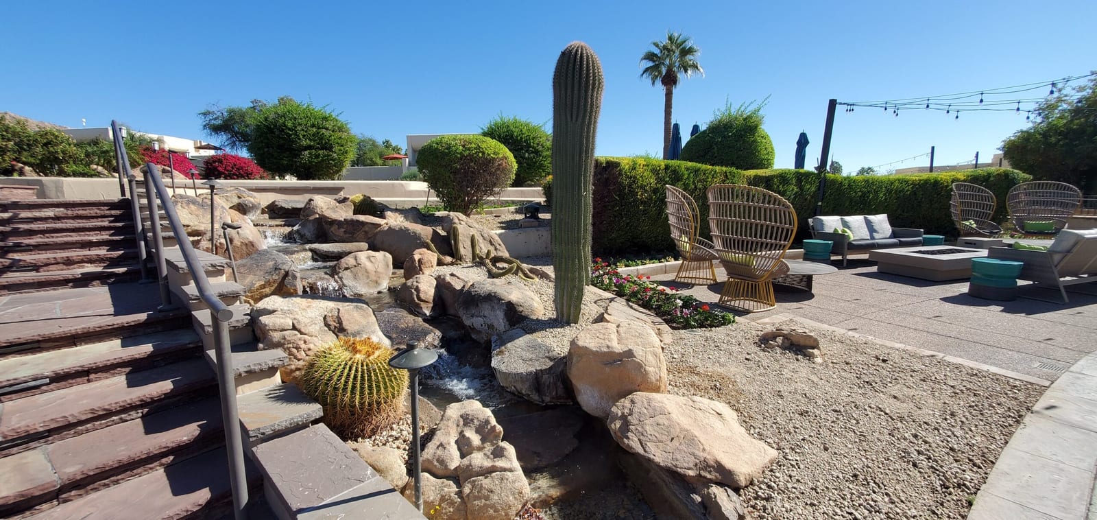 Serene waterfall stream and boulder garden bordering the outdoor patio lounge. Desert waterfall stream flowing past wicker lounge chairs and palm trees